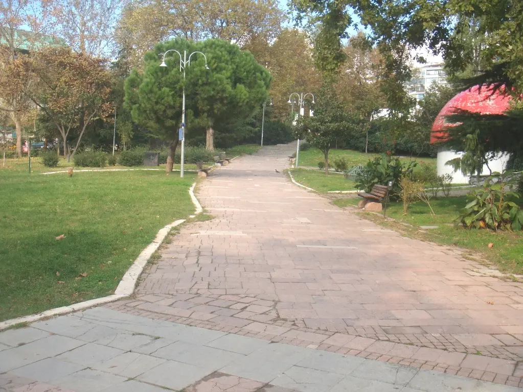 Maçka Demokrasi Parkı'nda kıvrılan pembe taş kaplama yürüyüş yolu; çimenlik, bank, fenerler, fıstık çamı ve kırmızı mantar çatılı büfe.