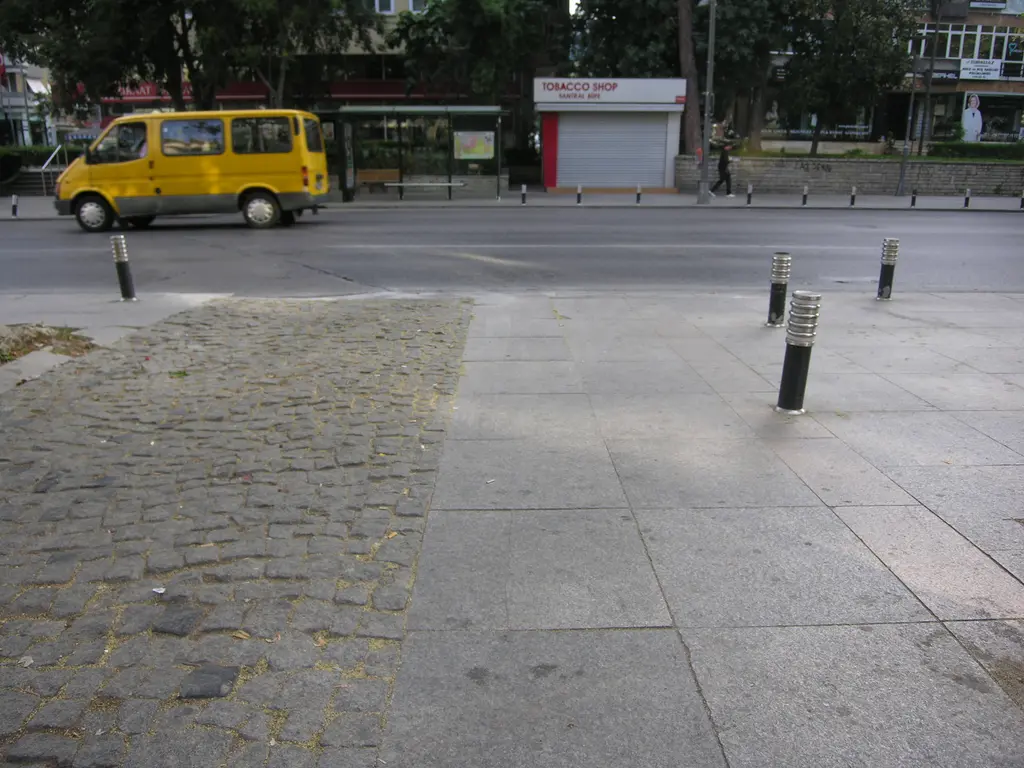Bağdat Caddesi'nde sabah; geniş taş kaldırım, demir direkler, karşı kaldırımda sarı dolmuş ve ağaçlar.