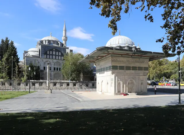 Tophane Meydanı'nda Tophane Çeşmesi; geniş saçaklı kare mermer gövde, yeşil zeminli yazı kuşağı, arkada Kılıç Ali Paşa Camii'nin kubbesi ve minaresi.