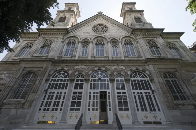 Taksim'de Aya Triada Kilisesi; iki çan kulesi arasında gül pencereli alınlık, iki katlı kemerli revak, açık renkli taş cephe ve mavi gökyüzü.
