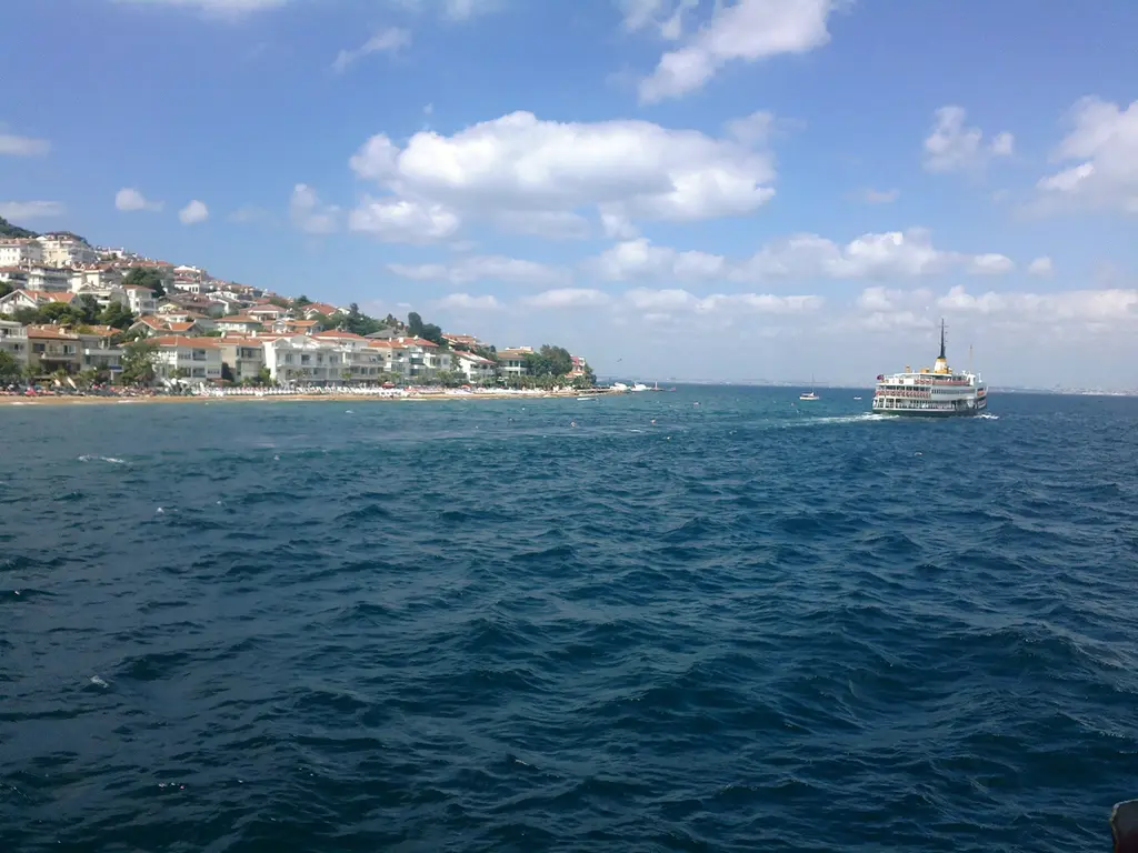 Adalar'da denizden bir ada; yamaca yayılan beyaz evler, dalgalı mavi deniz ve kıyıya yaklaşan Şehir Hatları vapuru.