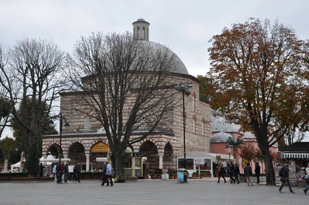 Hamamın taş gövdesi ve üstündeki büyük kubbe; altta kemerli revak, çevresinde çıplak ağaçlar ve önünden geçen birkaç kişi.