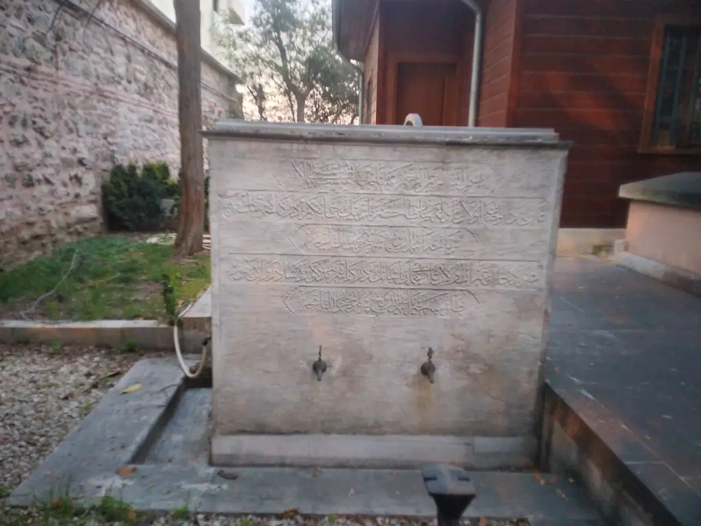 Cihangir Camii'nin avlusunda mermer çeşme; yazılı yüzey, iki musluk, taş zemin ve yanında ahşap kapılı yapı.