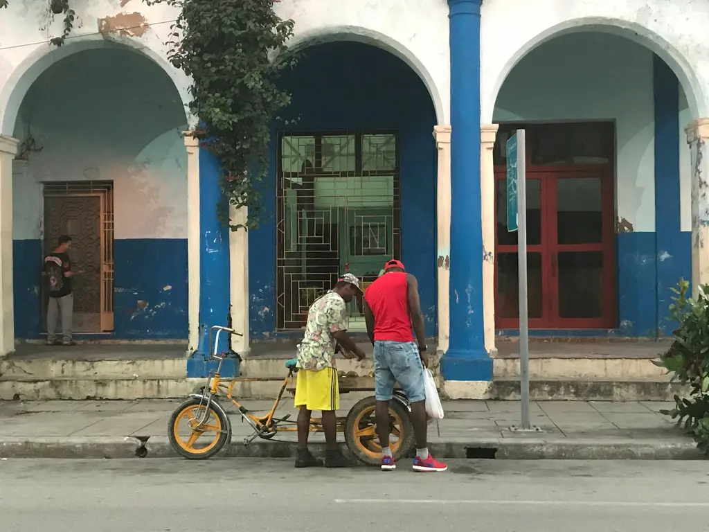 Camagüey'de mavi beyaz boyalı kemerli arkadlı bir binanın önünde sarı bisiklet taksi; yanında duran iki adam, kaldırımda tabela direği ve boş asfalt yol.
