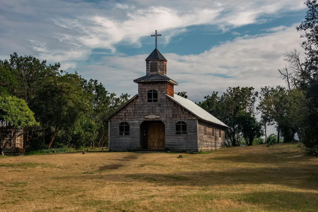 Chiloé'de ahşap kilise; tahta pul kaplamalı cephe, haçlı çan kulesi ve çevresini saran ağaçlarla çimenlik.