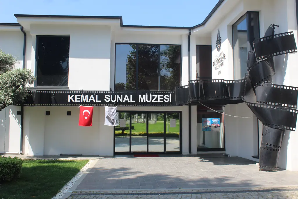 Göztepe Parkı'nda Kemal Sunal Müzesi; beyaz iki katlı bina, girişte film şeridi biçimli siyah heykel ve bayrak.