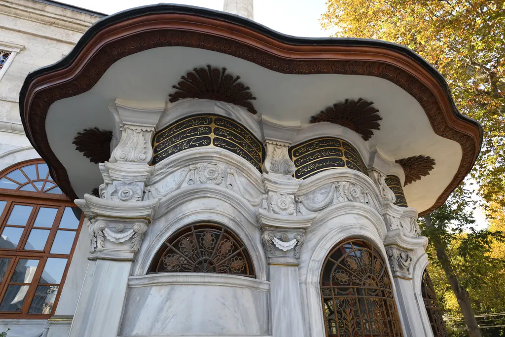 Nusretiye Camii'nin sebili; geniş dalgalı ahşap saçak, yaldızlı demir şebekeli kemerli pencereler ve kıvrımlı mermer cephe.