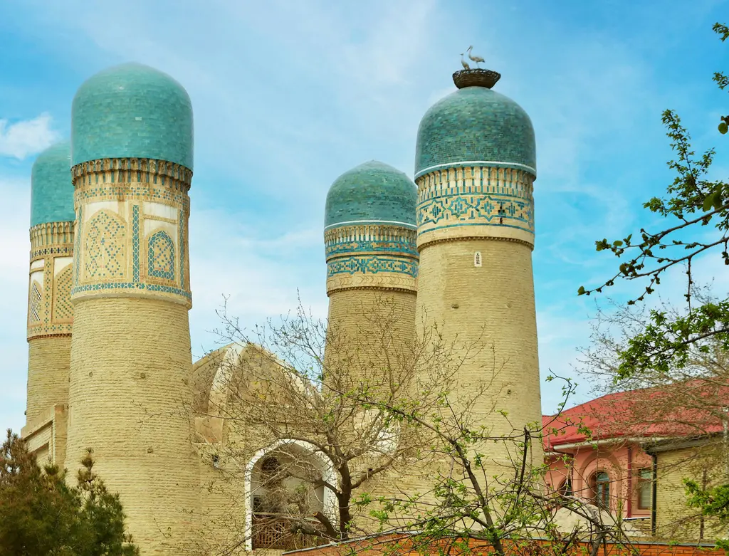 Buhara'da Miri Arab Medresesi'nin iki mavi kubbesi ve süslü portali; önünde meydan ve gezenler.