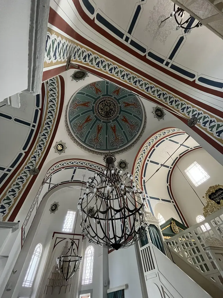 Atik Mustafa Paşa Camii'nin iç mekânında yukarı bakış; yeşil zeminli kubbe göbeği, kırmızı lacivert şeritli kemerler, hat madalyonları, dövme demir avize ve beyaz ahşap mahfil.