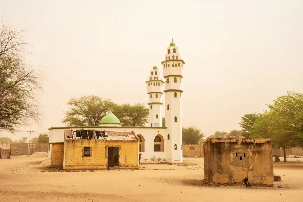 Senegal'de Sahel kuşağında beyaz badanalı bir cami; yeşil kubbesi ve iki minaresiyle, toz pusuyla örtülü gökyüzü altında kumlu bir meydanda.