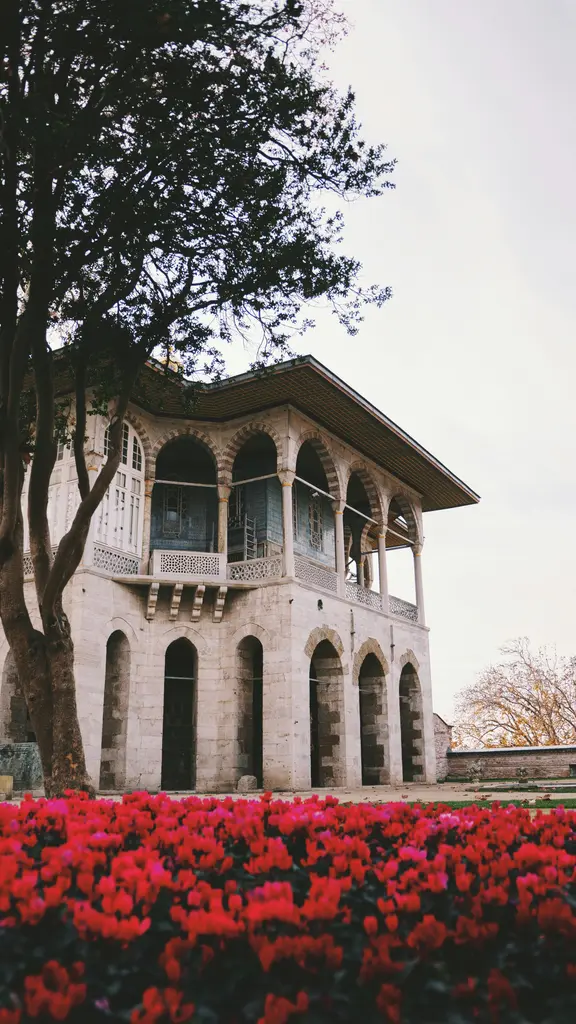 Topkapı Sarayı bahçesinde beyaz bir köşk; sütunlu cumbası, geniş saçağı, yanındaki ağaç ve önünde kırmızı lalelerle dolu tarhlar.