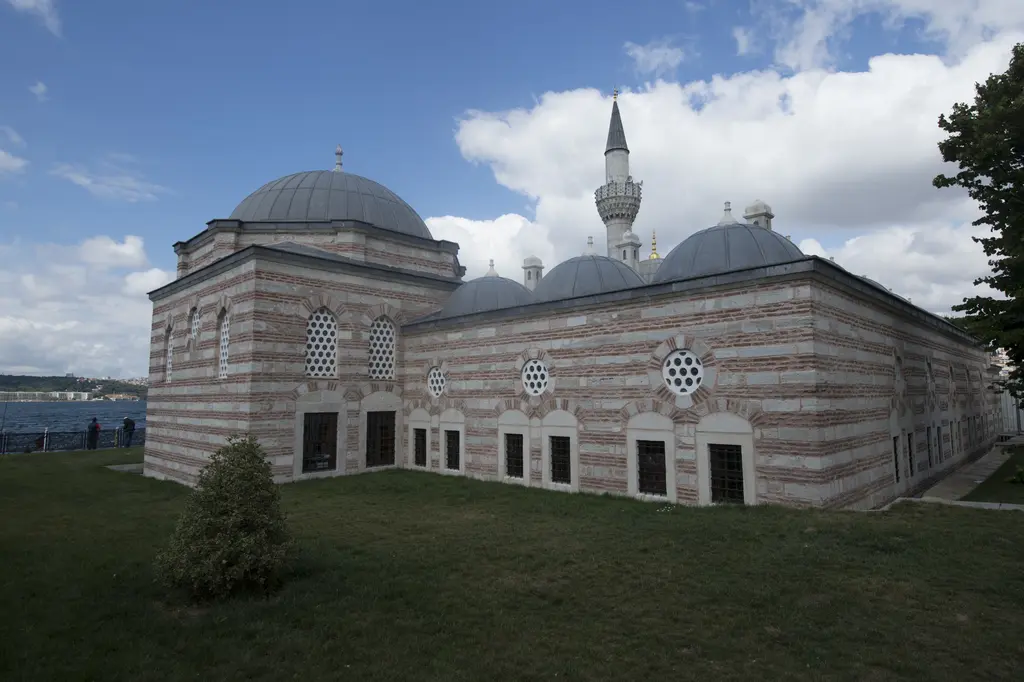 Şemsi Paşa Külliyesi; taş ve tuğla sıralı medrese duvarı, küçük kubbeler, arkada minare ve çimenlik.