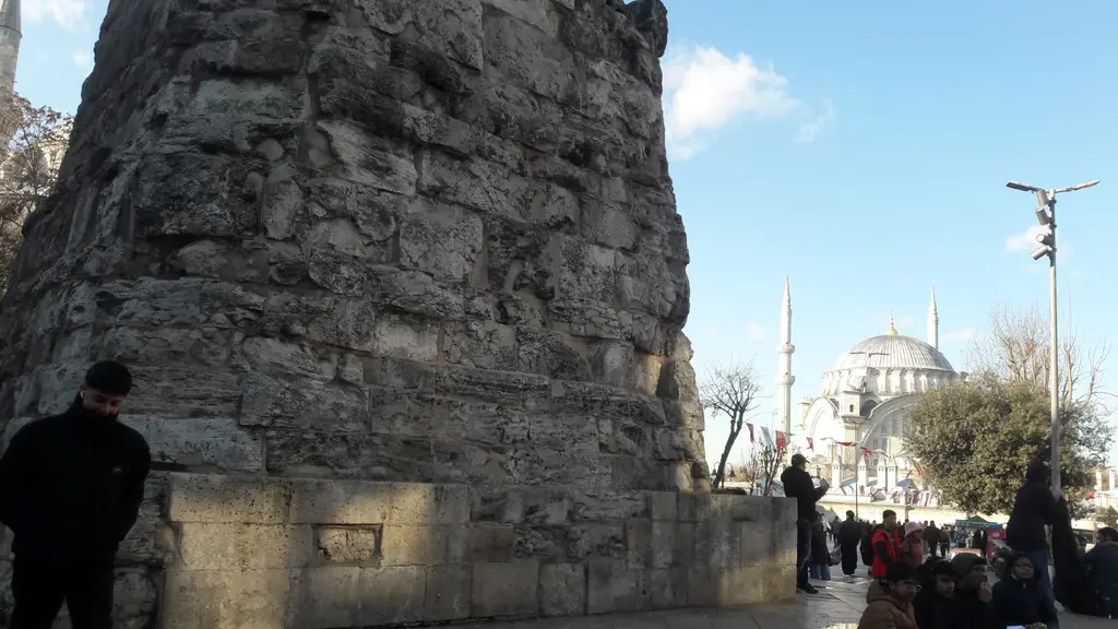 Çemberlitaş'ta taş kaidenin yakın planı; iri bloklardan örülü yüzey, kenarına dayanmış bir adam ve arkada kubbesiyle bir cami.