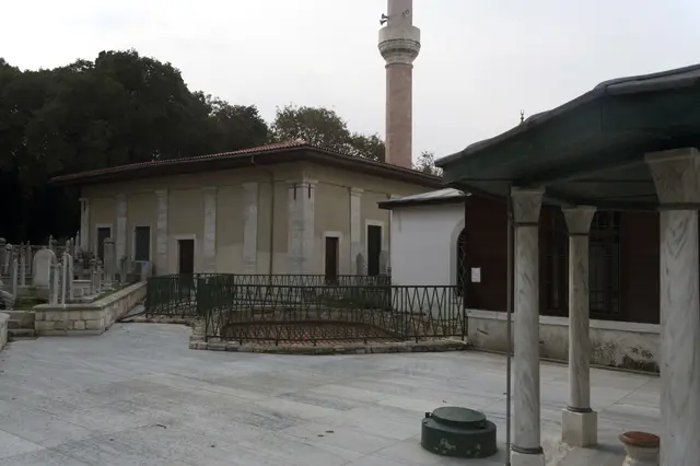 Merkez Efendi Camii'nin avlusu; ahşap direkli son cemaat revağı, kurşun kaplı çatı, solda tek şerefeli minare ve avluda eski mezar taşları.