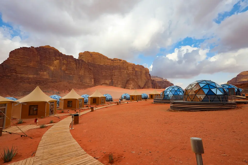 Wadi Rum'da kızıl kum üzerine kurulmuş çöl kampı; şeffaf kubbeli ve beyaz çadırlar, aralarındaki ahşap yürüme yolu ve arkada yükselen kayalık dağlar.