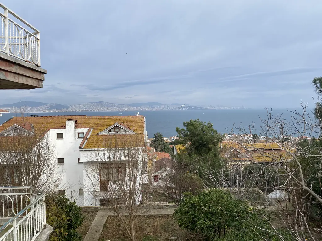 Kınalıada'da manzara; beyaz bir ada evinin çatısı, balkon korkulukları ve arkada Marmara ve Anadolu yakası.