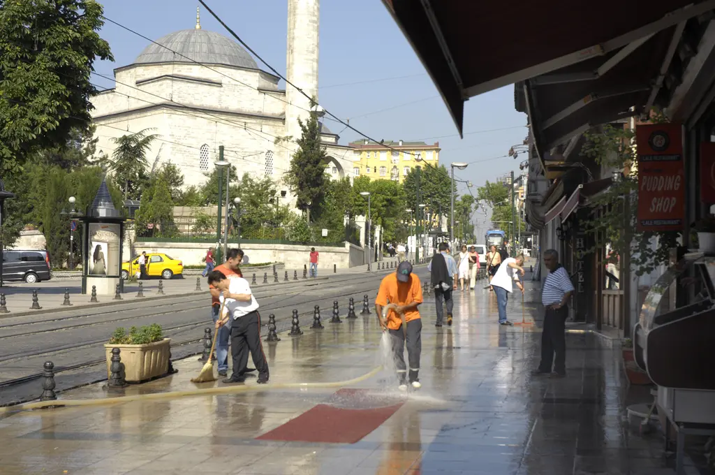 Yağmur sonrası Divanyolu; ıslak kaldırımı yıkayan temizlik görevlileri, bir tramvay durağı ve arkada Firuz Ağa Camii'nin kubbesi ile minaresi.