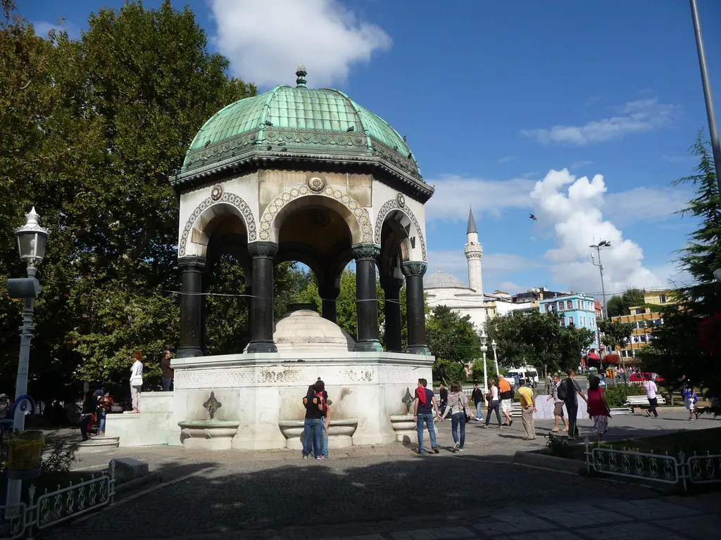Sultanahmet Meydanı'nda Alman Çeşmesi; yeşil bakır kubbe, koyu sütunlar, beyaz mermer kaide, çevresinde yürüyen insanlar ve uzakta bir minare.