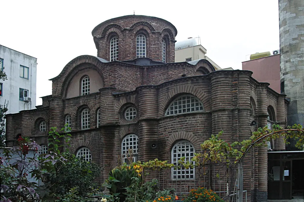 Bodrum Camii'nin kuzeybatı cephesi bahçeden; bordo yapraklı çalılar, sarı kanna çiçekleri, sağda asma çardağı ve minare kaidesinin taş yüzeyi.