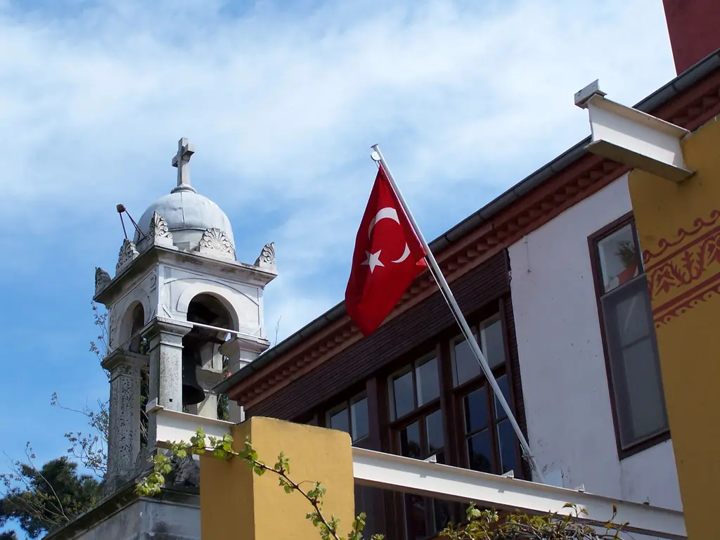 Büyükada'da Aya Yorgi Kilisesi; çan kulesi, bayrak, ahşap çıkmalı yapı ve kiliseye çıkan yolda ziyaretçiler.