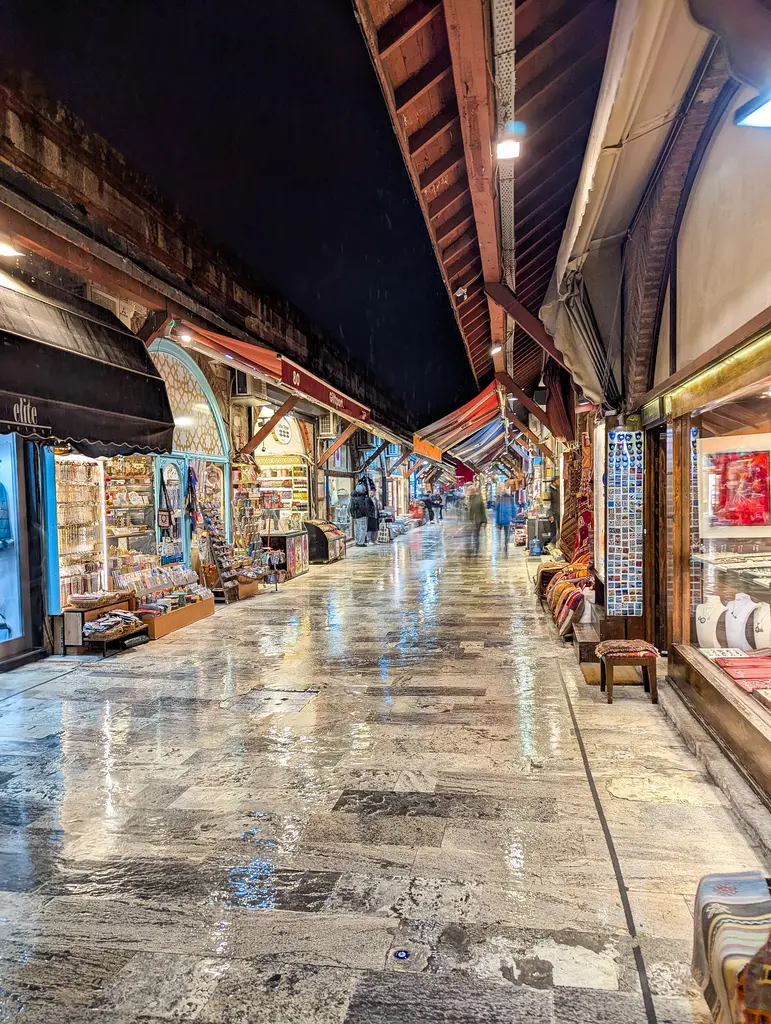 Akşam Arasta Çarşısı; ışıklı dükkân vitrinleri, parlayan mermer zemin, yukarıda koyu tenteler ve sokağın sonuna uzanan dükkân sırası.