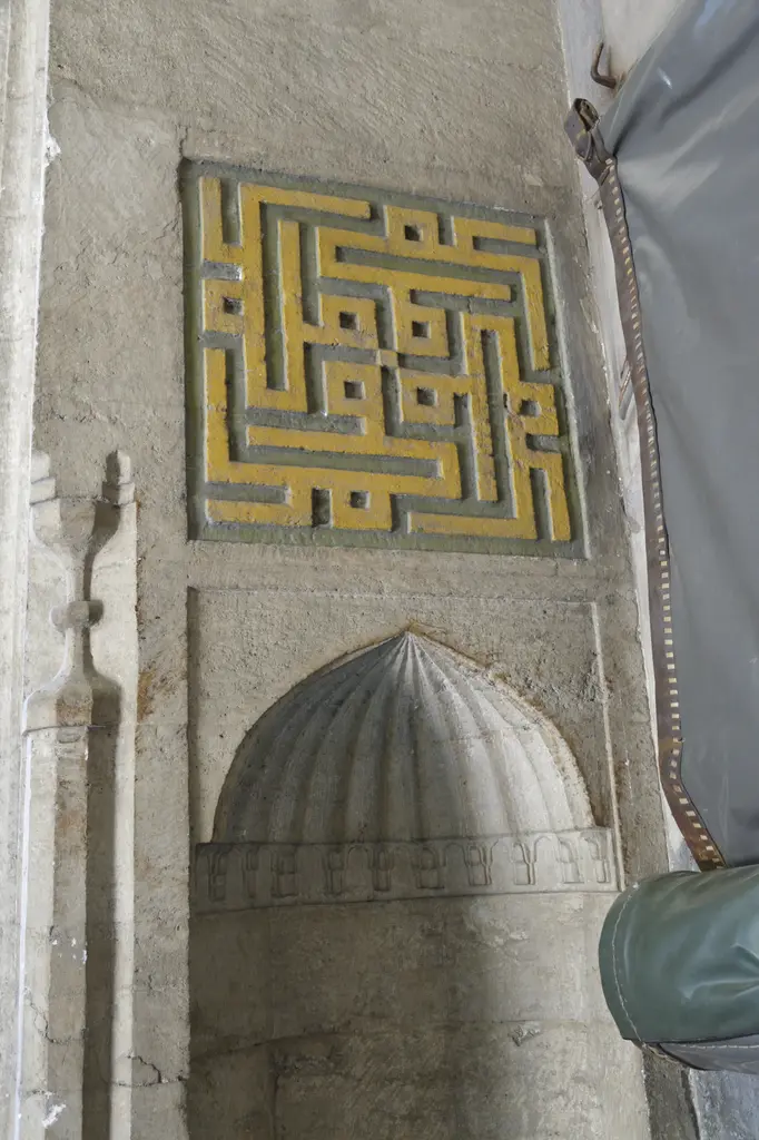 Haseki Sultan Camii'nde mihrabiye nişinin üstünde sarı ve siyah köşeli kufi yazı panosu, yanda perde ve taş duvar.
