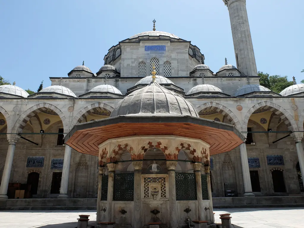 Sokullu Mehmed Paşa Camii'nin şadırvanı; sütunların taşıdığı konik çatı, mermer şebekeli gövde ve arkasında caminin revaklı cephesi.