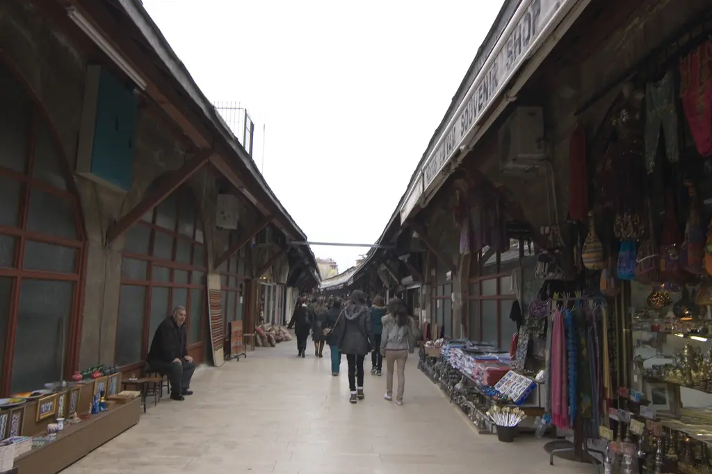 Arasta Çarşısı'nda uzun perspektif; iki yanda ahşap sundurmalar, kemerli dükkânlar, önlerde sergilenen kilimler ve oturan bir esnaf.