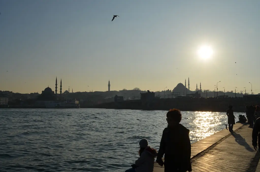 Karaköy rıhtımında gün batımı; kıyıda oturan insanların siluetleri, parlayan su ve karşıda tarihi yarımadanın camileri.