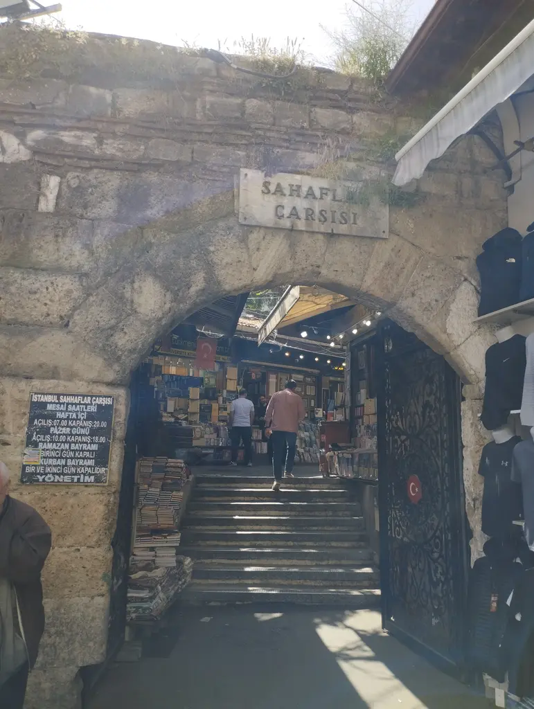 Sahaflar Çarşısı'nın kemerli geçidi içeriden; kaba taş kemer, üstünde mermer levha, sağda ay yıldızlı dökme demir kanat, altta çarşıya çıkan taş merdiven ve iki yanında kitap yığınları.