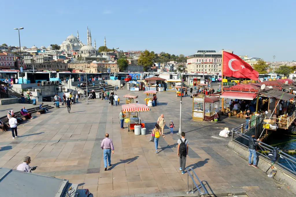 Eminönü meydanı ve kıyısı; geniş taş meydan, seyyar simitçi arabaları, balık ekmek tekneleri, Türk bayrağı ve tepede Süleymaniye Camii.
