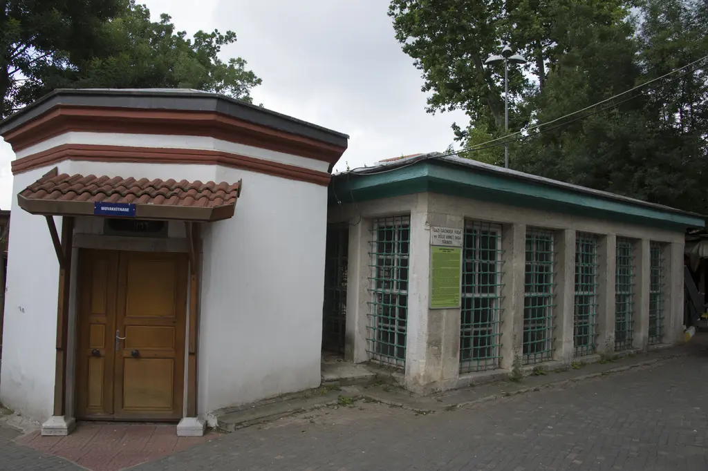 İskender Paşa Türbesi; beyaz sıvalı köşeli yapı, ahşap kapı, saçaklı giriş ve yanında yeşil pencereli bina.