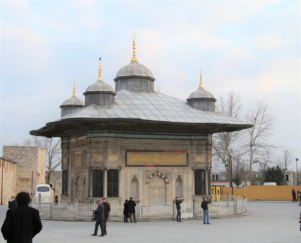 Meydanda çeşme; önünden geçen ziyaretçiler, çevresindeki beyaz parmaklık, arkada saray duvarı ve ağaçlar.