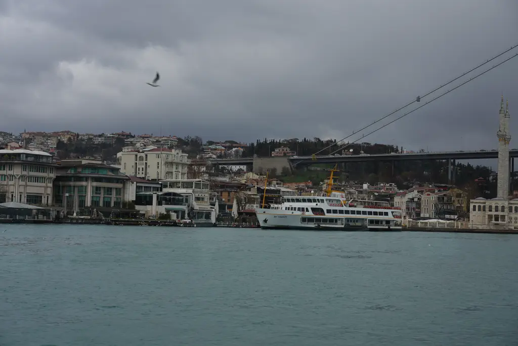 Denizden Ortaköy kıyısı; bulutlu gökyüzü altında köprünün ayağı, kıyı boyunca binalar, iskele ve suda ilerleyen tekneler.