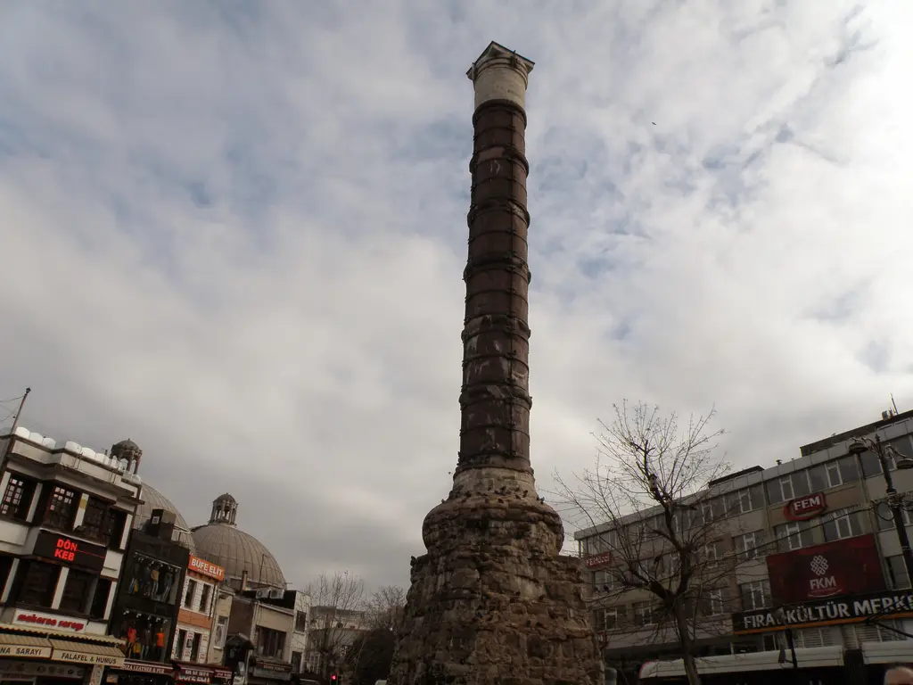 Çemberlitaş'ta kapalı havada sütun; gri bulutlu gökyüzü, koyu gövde ve çevresini saran cadde binaları ile dükkân tenteleri.