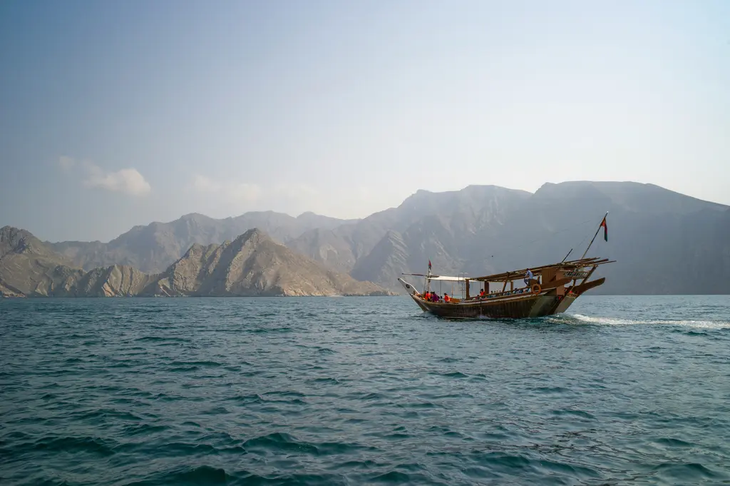 Musandam'da koyu mavi suda yelken direkli ahşap dhow teknesi; arkasında çıplak gri-kahve kayalık yamaçlar ve puslu gökyüzü.