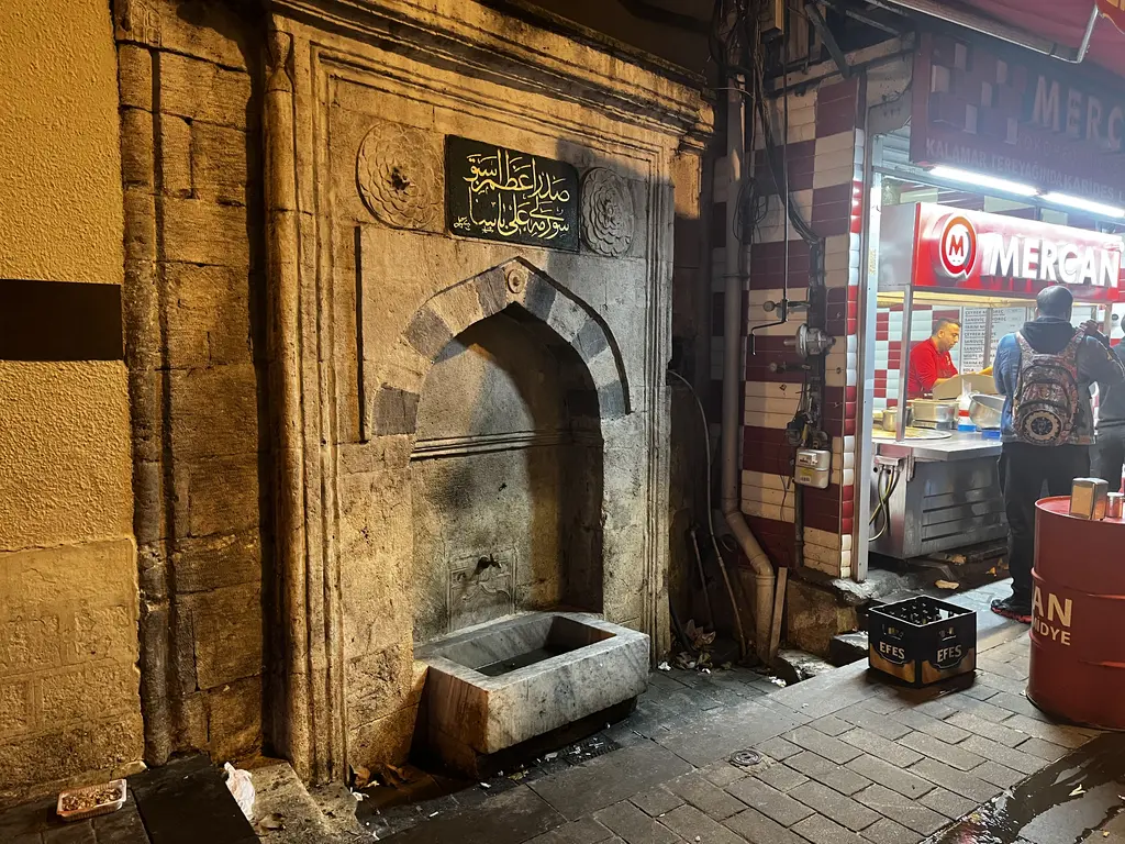Kadıköy Çarşısı'nda Sürmeli Ali Paşa Çeşmesi; kemerli taş niş, yalak, Osmanlıca kitabe ve yanda dükkân.