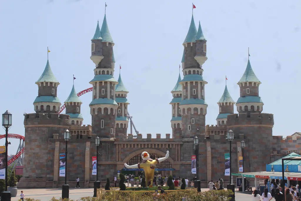 Vialand'de karşıdan görülen şato; turkuaz külahlı yüksek kuleler, mazgallı duvarlar, önde altın maskot heykeli, geniş meydan ve ziyaretçiler.