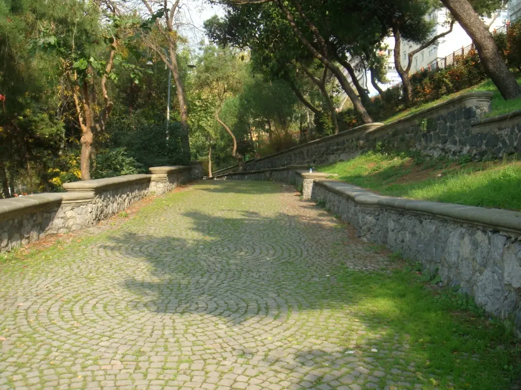 Maçka Demokrasi Parkı'nda yelpaze desenli parke taşı yol; iki yanında taş istinat duvarları, çimenli şevler ve üstte ağaçlar.