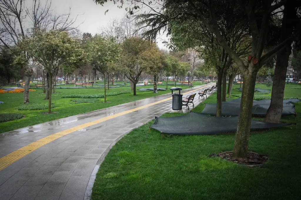Göztepe Parkı'nda yürüyüş yolu; yağmurla ıslanmış yol, sarı hissedilebilir şerit, ağaçlar, banklar ve çimenlik.