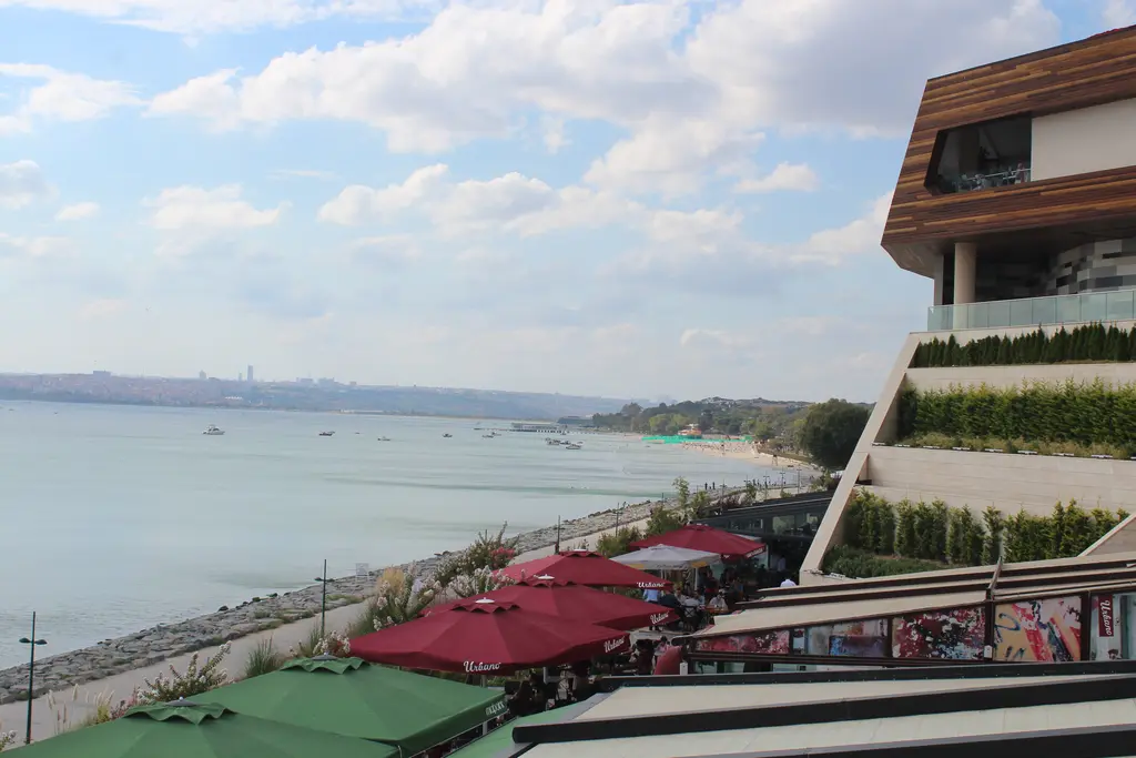 Aqua Florya'nın teraslarından Florya sahili; kırmızı şemsiyeli kafe terası, kıyı boyunca uzanan yol, sakin deniz ve uzakta kıyı şeridi.