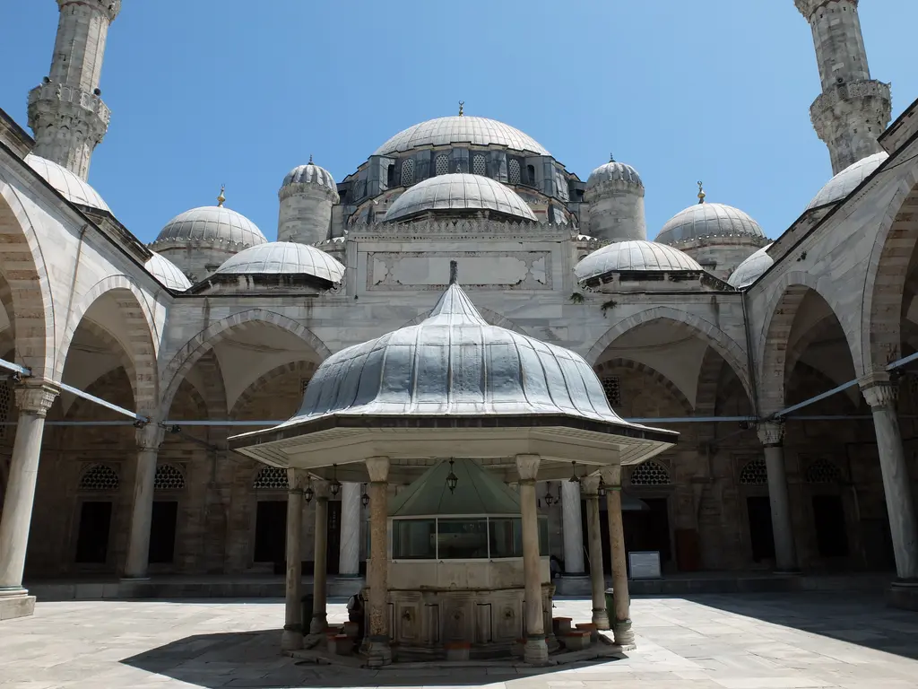 Şehzade Camii'nin avlusu alttan; ortada kurşun külahlı şadırvan, çevresinde revak kemerleri, üstte kubbeler ile minare kaideleri ve koyu mavi gökyüzü.