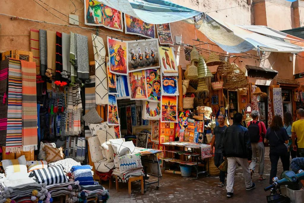 Marakeş çarşısında (souk) bir sokak; asılı renkli fenerler, halılar, baharat tezgâhları ve kalabalık.