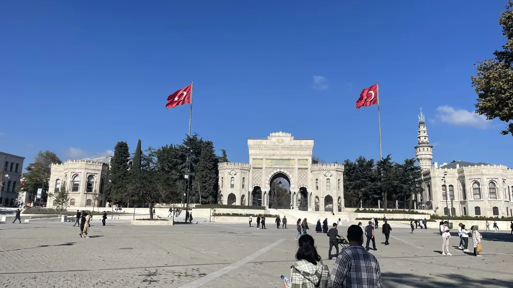 Beyazıt Meydanı'ndan üniversite kapısı ve kule; geniş taş döşeli meydan, bayrak direkleri, birkaç yaya ve arkada kulenin gövdesi.