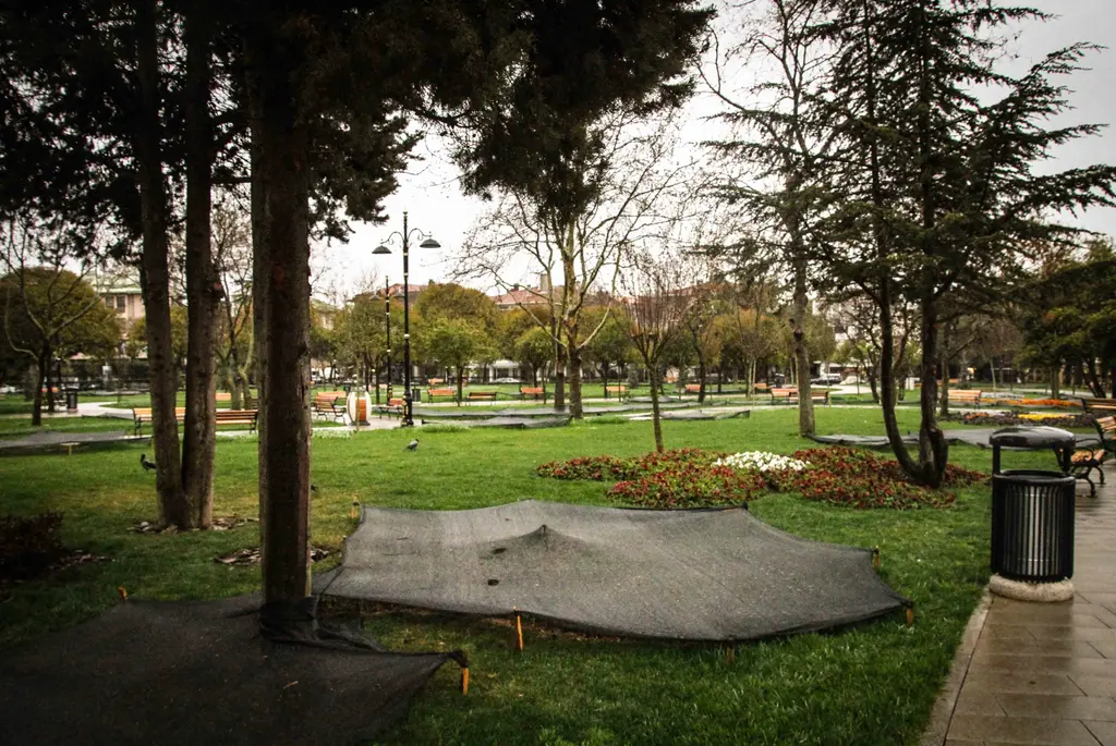 Göztepe Parkı'nda bahar başı; ağaçlar arasında banklar, gölgelik örtülü tarhlar, çöp kutusu ve taş kaplı yol.