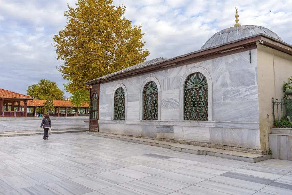 Sonbaharda türbe ve çevresi; sarı yapraklı ağaçlar, mermer cepheli türbe, arkada caminin kubbesi ve önde yürüyen bir ziyaretçi.