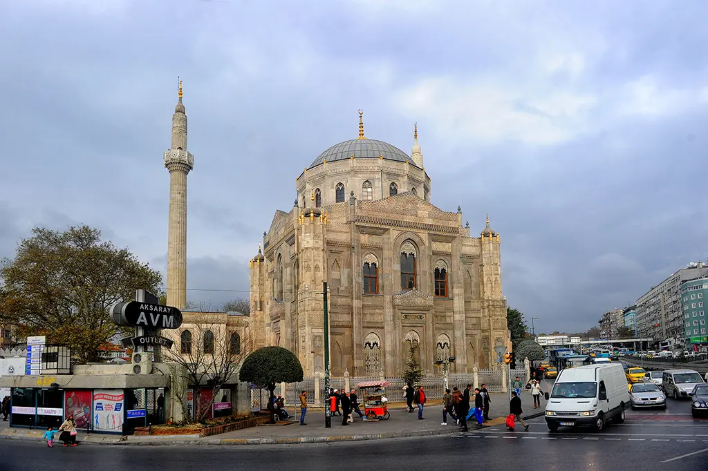 Aksaray kavşağından Pertevniyal Valide Sultan Camii; önde trafik ve kaldırımda yürüyenler, arkada minare, kubbe ve güneşli cephe.