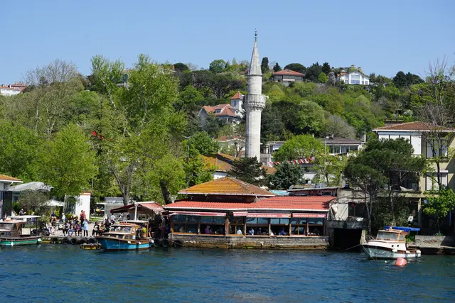 Kanlıca'ya denizden bakış; kıyıda kırmızı tenteli lokanta, tekneler, ağaçlar arasında yükselen ince minare ve sırttaki evler.