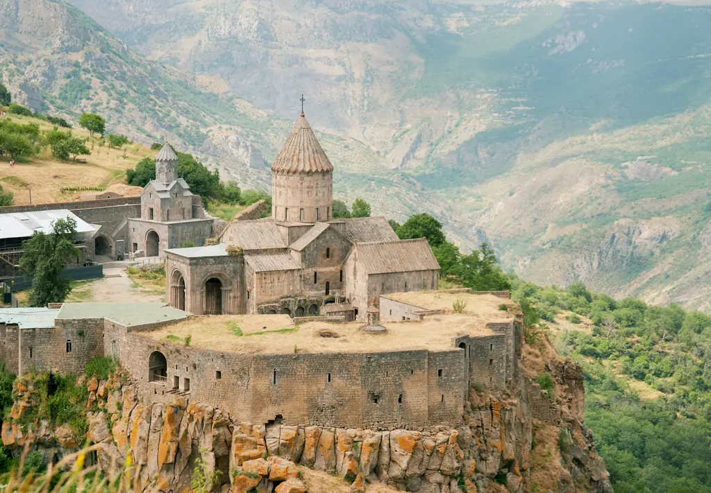 Ermenistan'da derin bir vadinin kenarındaki uçurumda yükselen Tatev Manastırı; konik çatılı taş kilise ve çevresini saran sur duvarları, arkada sisli yeşil dağlar.