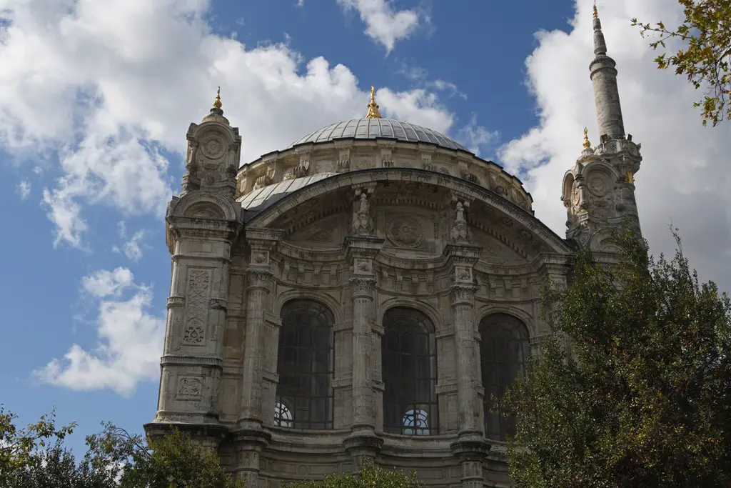 Ortaköy Camii'nin cephesi; yivli sütunlar, kemerli pencereler, oymalı alınlık, yanda ince minare ve beyaz bulutlar.